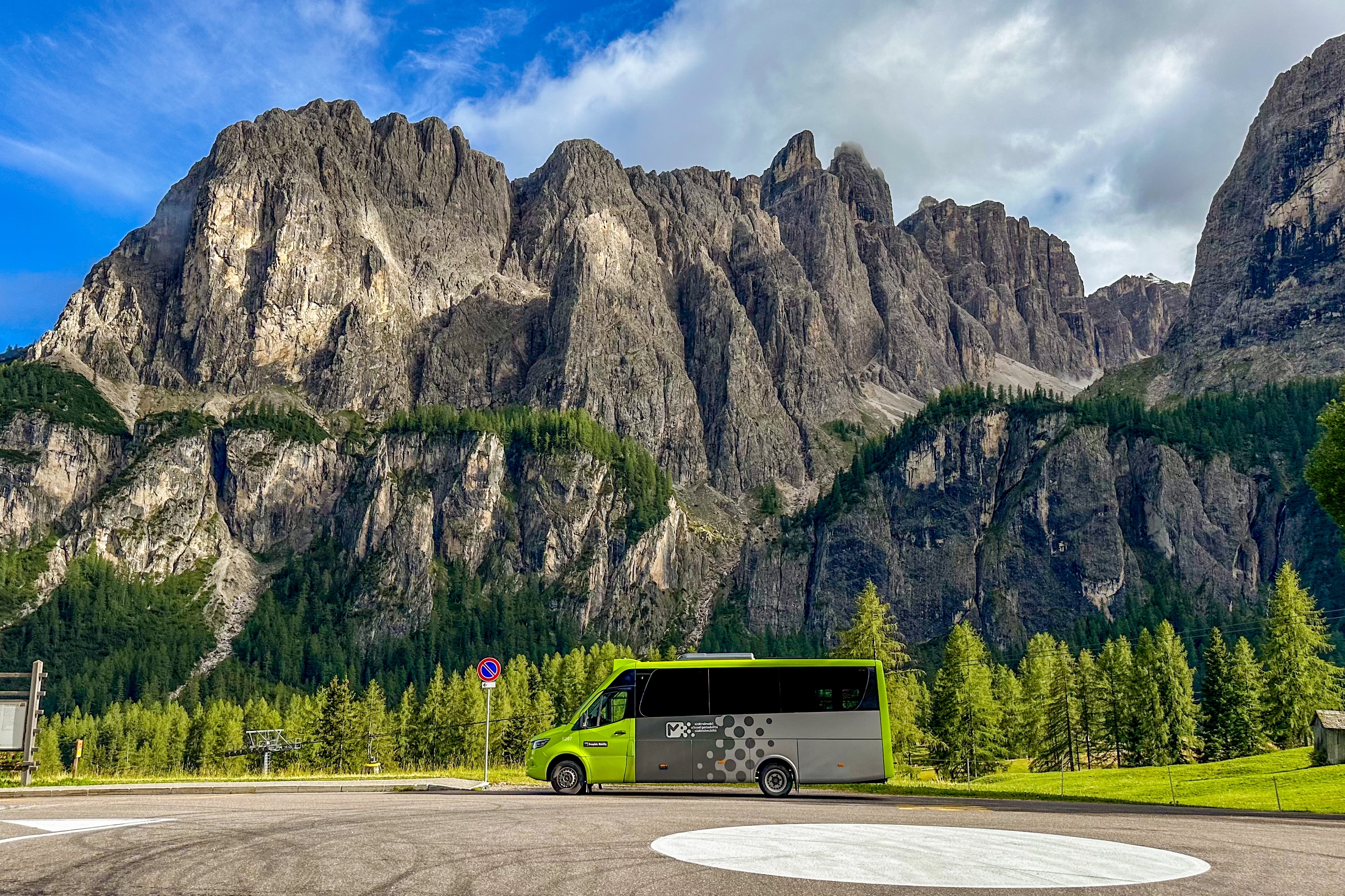  Green and silver minibus parked by roadside with majestic Dolomite peaks and forest landscape in the background under clear sky